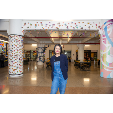 A person stands confidently in a bright, modern indoor space decorated with colorful hanging ornaments and wall art, with tables and chairs in the background.