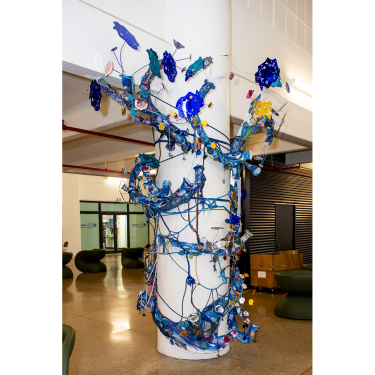 A white column is wrapped in a colorful, intricate art installation with blue, yellow, and purple elements, weaving vines, flowers, and abstract shapes, located inside a modern building with glass doors and seating nearby.