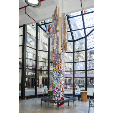 A tall, colorful abstract art installation hangs from the ceiling inside a bright, spacious atrium with large windows, modern furniture, and industrial architecture visible outside.