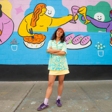 Hyesu Lee in colorful clothes stands smiling with arms crossed in front of a bright mural showing cartoon people eating and toasting with food and drinks. The mural features bold, cheerful colors and playful designs.