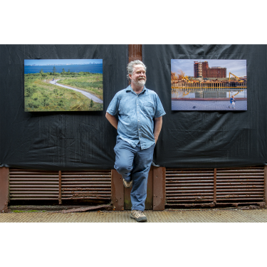 A man with gray hair and a beard, dressed in a blue shirt and pants, stands against a black backdrop with two landscape photographs displayed on either side of him.