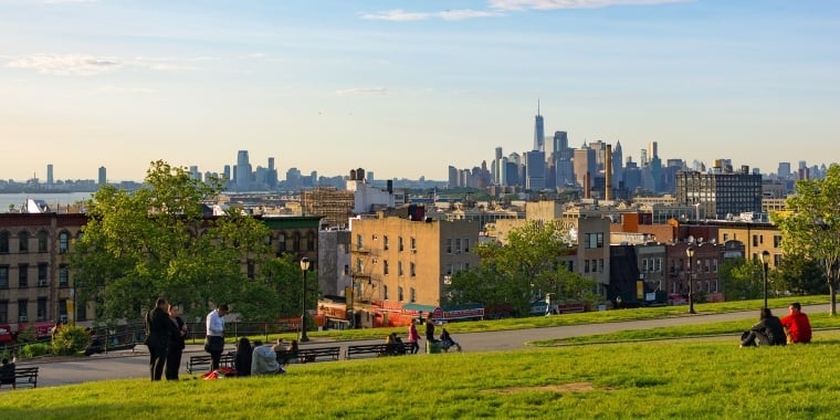 A grassy hill in Sunset Park, Brooklyn overlooking the New York City skyline at sunset. People sit on the grass and benches, enjoying the view. The sky is partly cloudy, with the sun casting warm light on the scene. Tall buildings are visible in the distance.