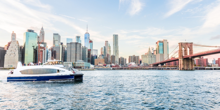 View of NYC Ferry with Lower Manhattan skyline and Brooklyn Bridge in the background at sunset