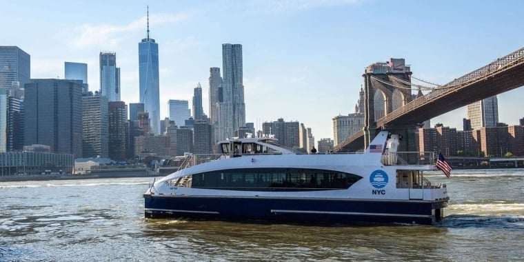 NYC Ferry sails on the river with the Manhattan skyline and Brooklyn Bridge in the background on a clear day.