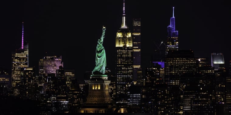 Night view of the New York City skyline featuring the illuminated Statue of Liberty in the foreground and skyscrapers in the background.