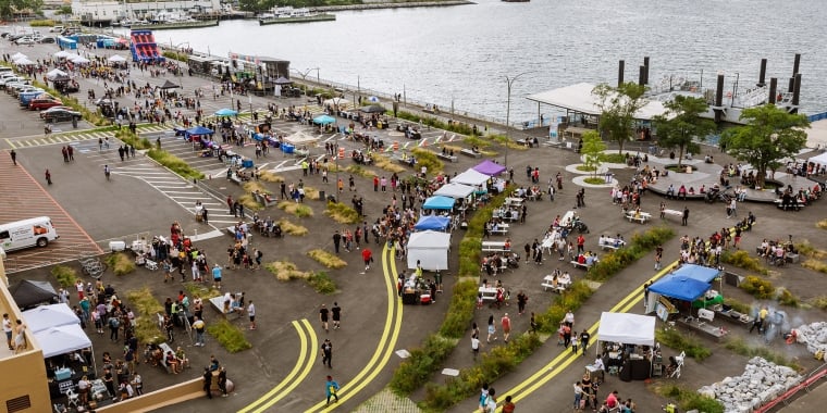 Aerial view of an outdoor food festival by the waterfront. Crowds of people gather around various food stalls and seating areas. The scene includes colorful tents and a view of the water with ships in the distance.