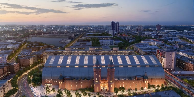 Aerial view of a large, illuminated Kingsbridge Armory building with two towers, surrounded by cityscape at dusk. The building features a glass-domed roof and is set against a cloudy sky with a pinkish hue. Streets and trees are visible around the structure.