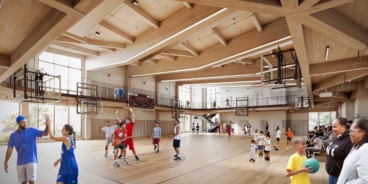 A large indoor basketball court with people playing various sports. Children and adults are engaging in activities like basketball and passing balls. Spectators are watching from a mezzanine level. The hall features wooden ceilings and large windows.