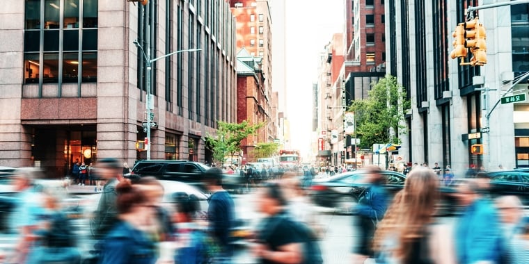 A busy city street scene with blurred motion capturing people crossing at a crosswalk. Tall buildings line both sides of the street, and the background reveals a mix of trees and urban architecture. Traffic lights are visible above the intersection.