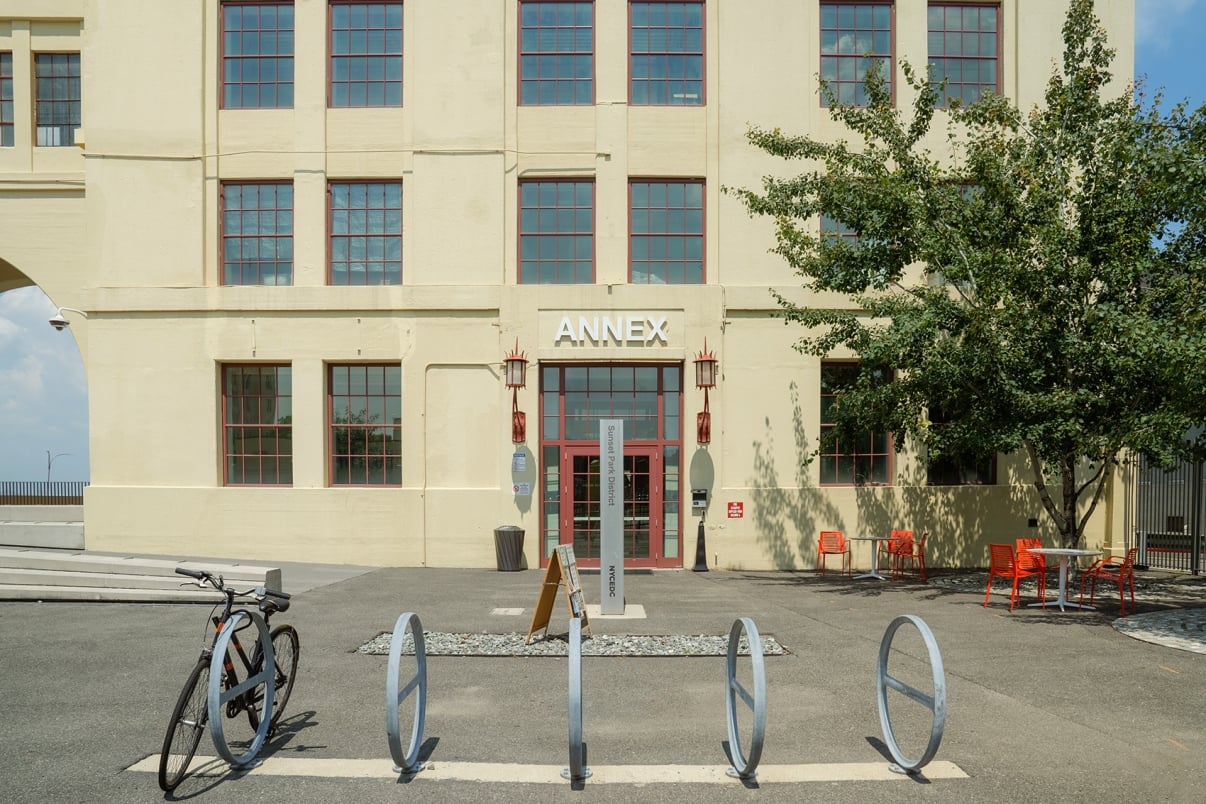 A large, beige building complex with multiple stories and arched windows. In the foreground, an open-air bridging structure connects parts of the building. The sign Building A is visible above an entrance with outdoor benches nearby.