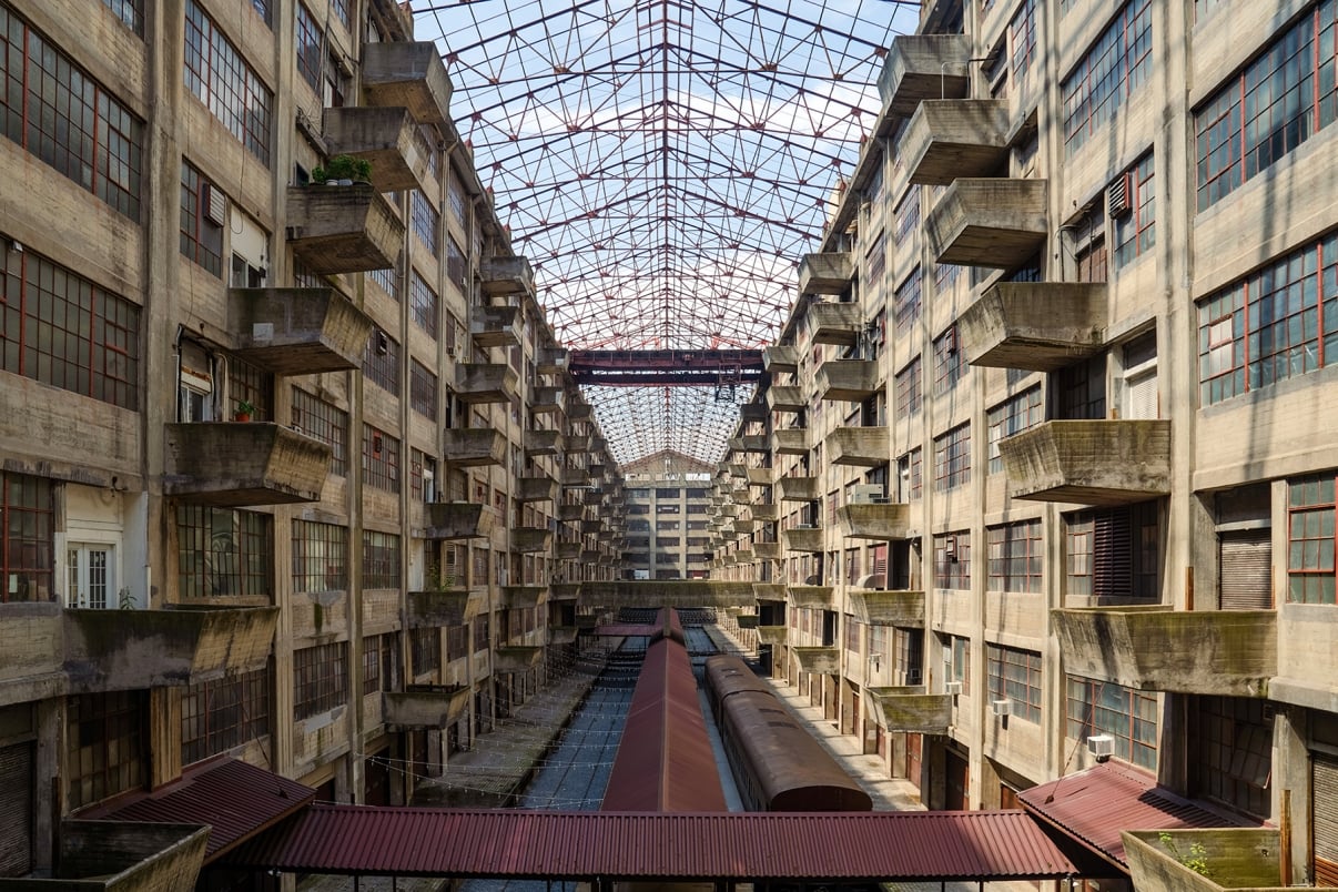 Interior of the Brooklyn Army Terminal Atrium with multiple floors and uniform balconies. Natural light filters through the metal ceiling, casting shadows. There is an old train is visible on the ground floor.