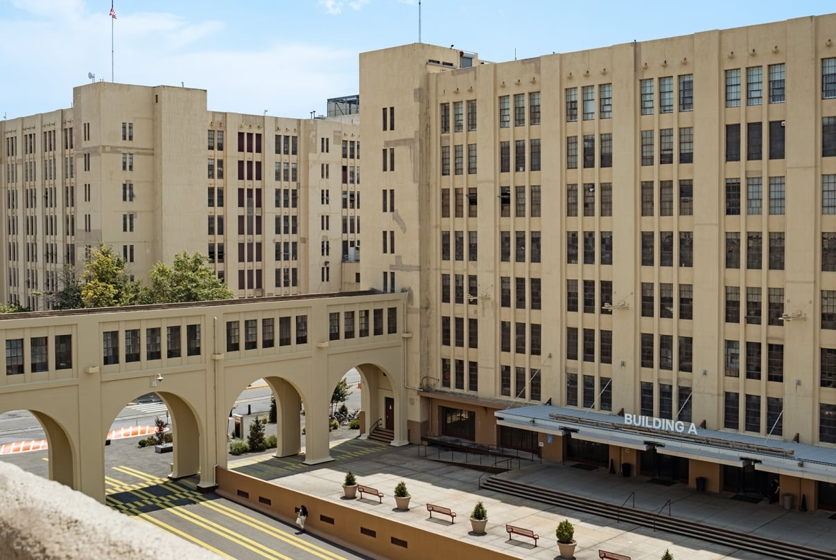 A large, beige building complex with multiple stories and arched windows. In the foreground, an open-air bridging structure connects parts of the building. The sign Building A is visible above an entrance with outdoor benches nearby.
