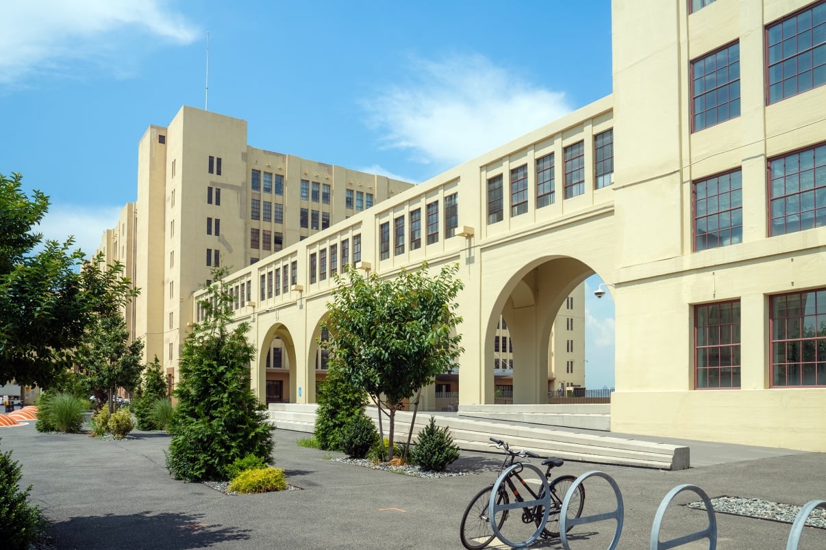 A large beige industrial building with tall windows and arches, set against a clear blue sky. The foreground features a small garden with trees and a bicycle parked on a paved area.