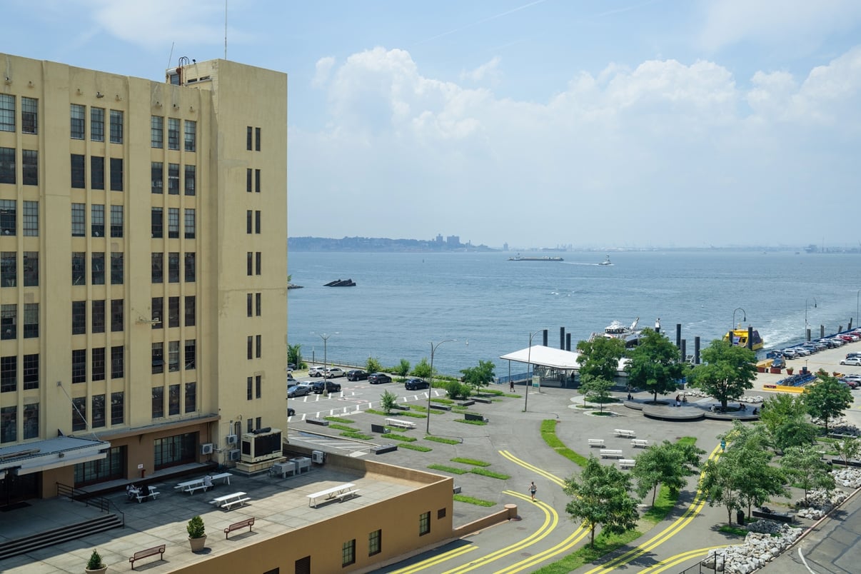  A coastal industrial area with a large beige building on the left, overlooking a body of water. There are parked cars and trees scattered around the paved area. In the background, ships are visible on the water under a cloudy sky.