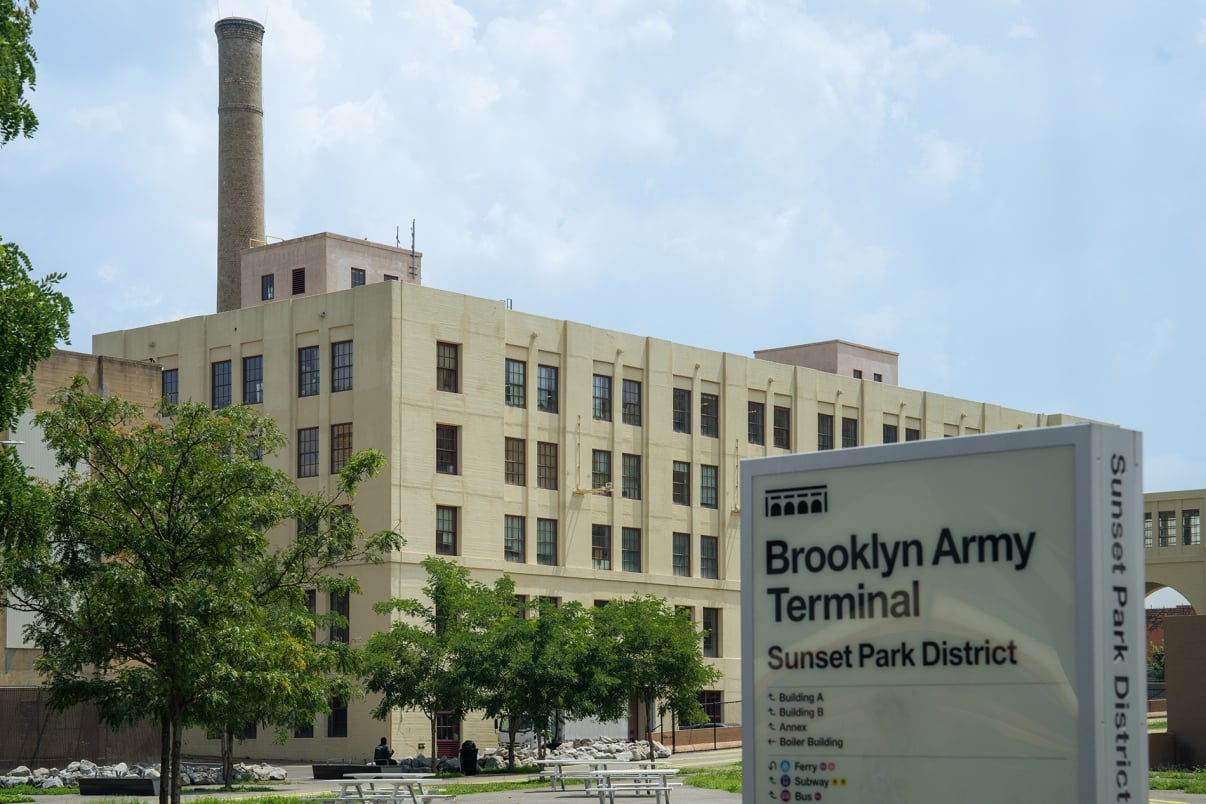 The image shows the Brooklyn Army Terminal building, a large beige industrial structure with a tall chimney. In the foreground, theres a sign reading Brooklyn Army Terminal, Sunset Park District and trees lining the entrance.