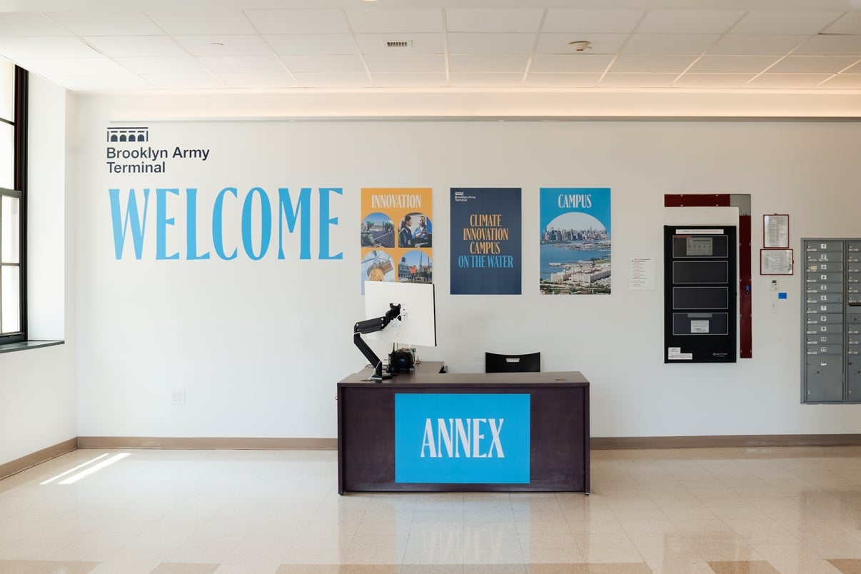  A welcome desk labeled Annex in an office area with posters on the wall above it. The posters display the words Innovation and Campus along with city imagery. A light fixture and wall-mounted utility panels are visible beside the desk.