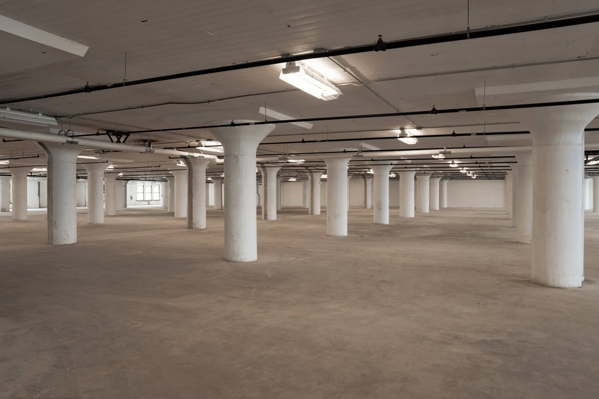 A large, empty warehouse interior with white columns evenly spaced throughout. The ceiling is exposed, showing pipes and fluorescent lights. The concrete floor is plain, and a window is visible in the background.