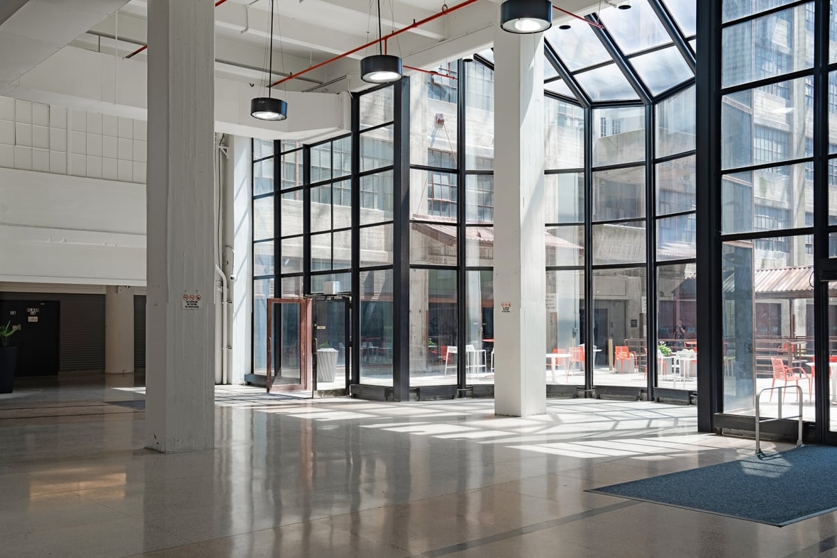  A spacious, sunlit atrium with large glass windows and a high ceiling. There are several tables and chairs outside, visible through the windows. Light reflections cast patterns on the polished floor.