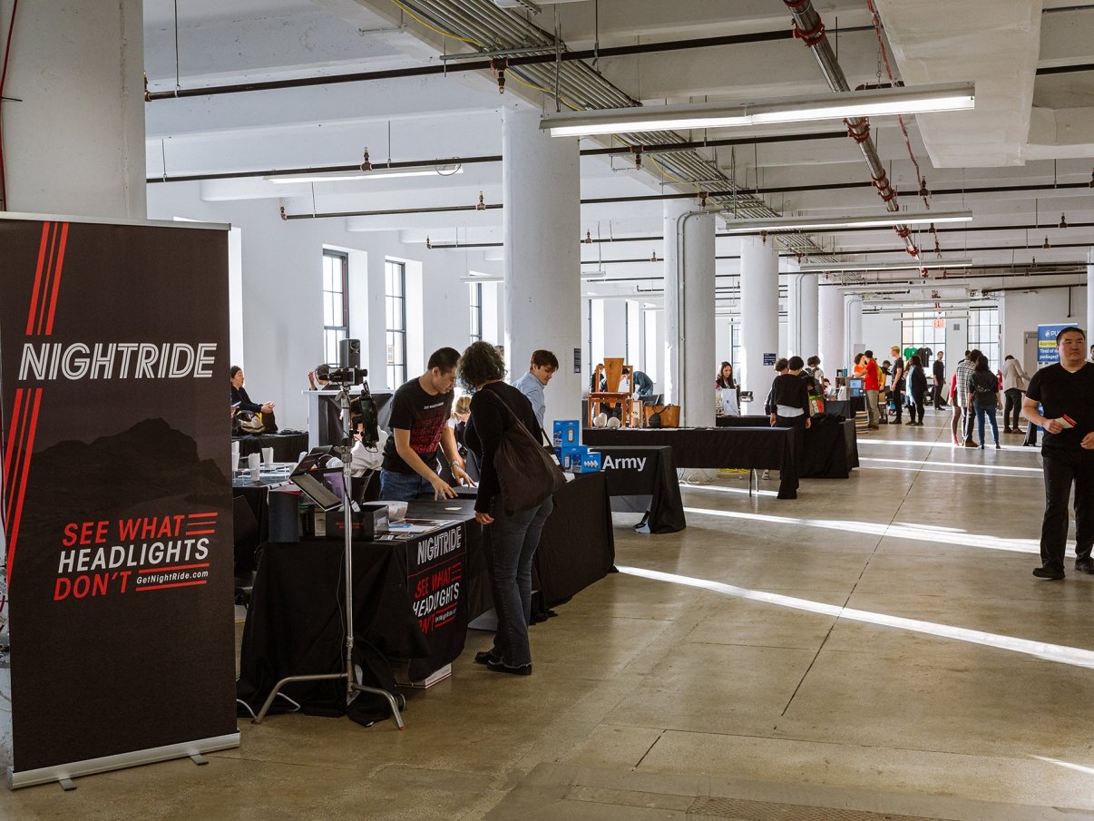 A spacious event hall with several booths and people interacting. A prominent booth displays the brand Nightride with the tagline See What Headlights Don&#039;t. Bright lighting from large windows illuminates the room.