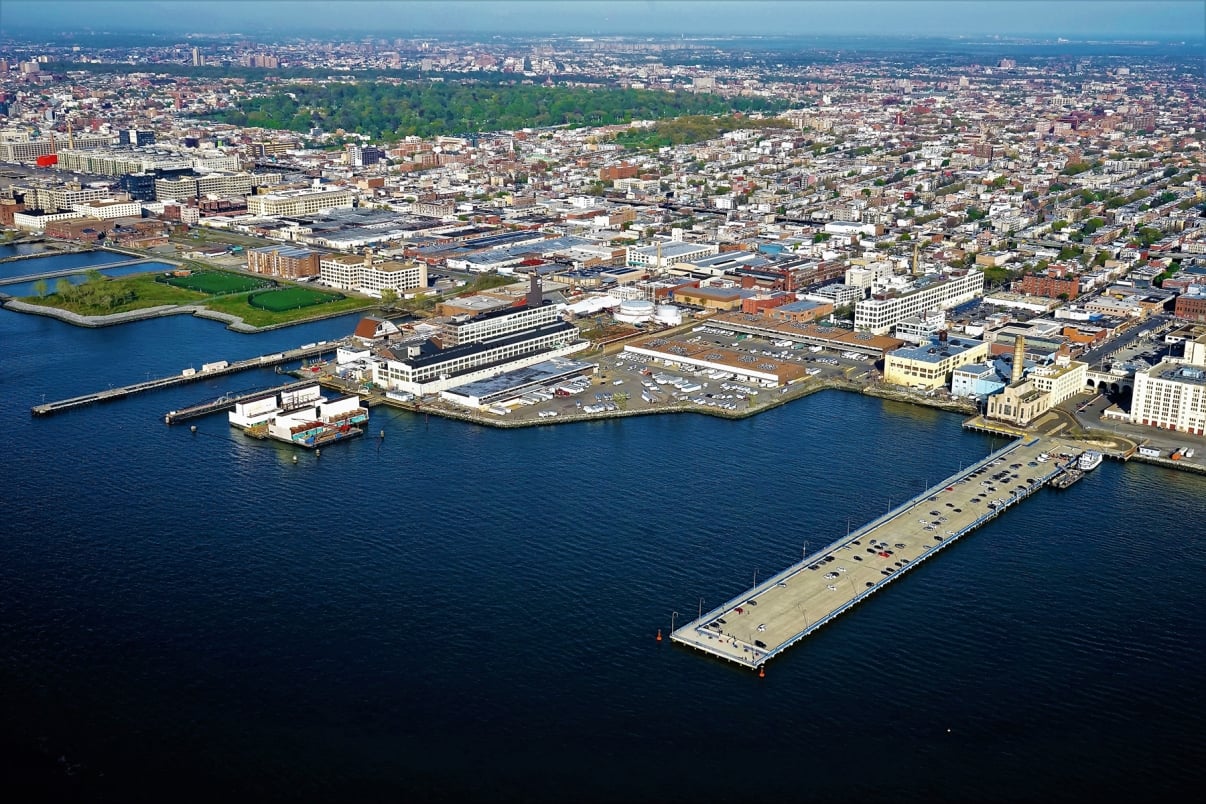 Aerial view of a Sunset Park and the Brooklyn waterfront showing a long pier extending into the water. The cityscape includes a mix of buildings, streets, and green areas. A body of water surrounds the pier, with urban development visible in the background.