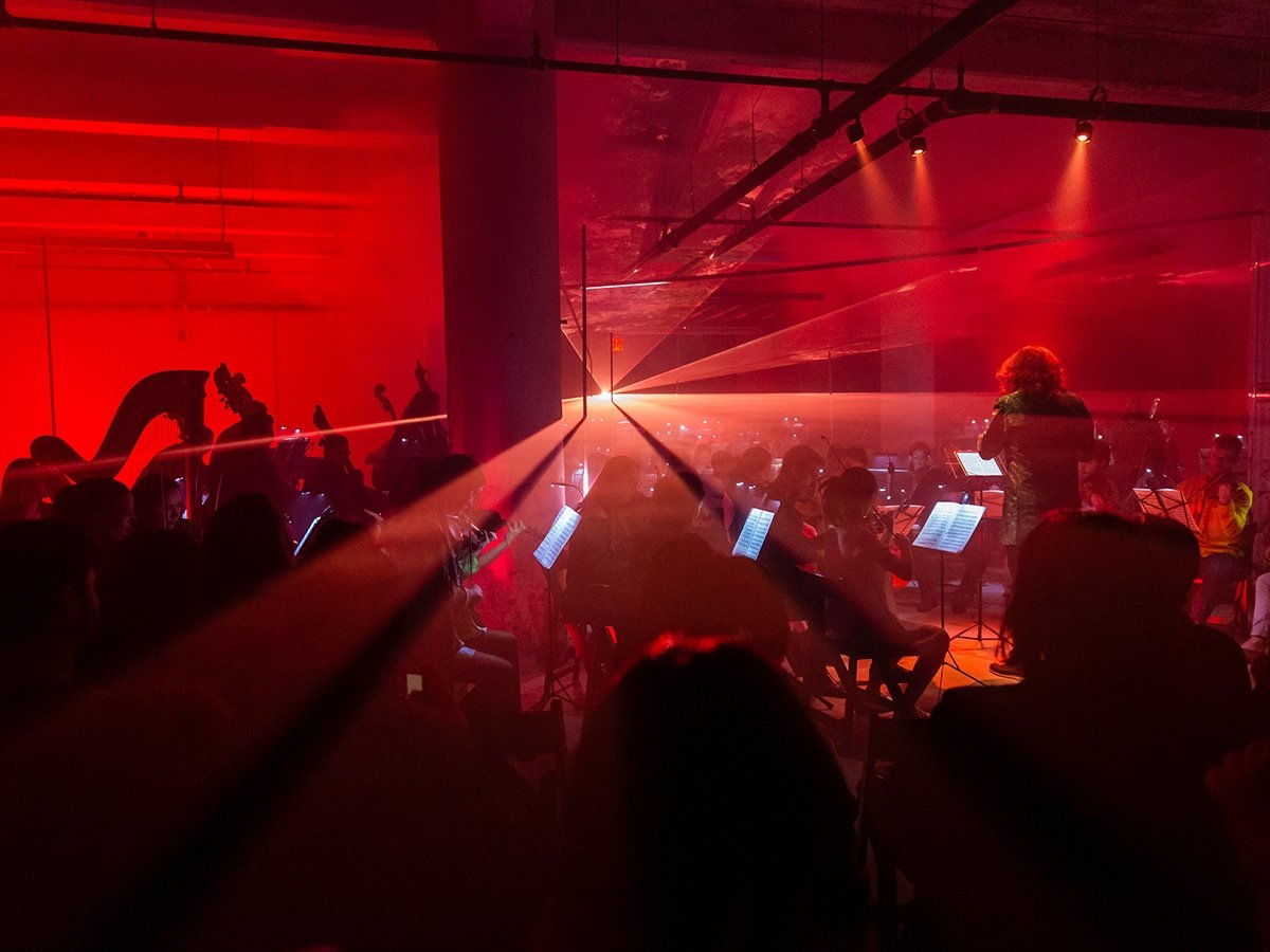 (direct upload) A vivid image of an orchestra performing under dramatic red lighting. Musicians play various instruments, and a conductor leads them. A bright light beam cuts through the scene, creating an intense and dynamic atmosphere.