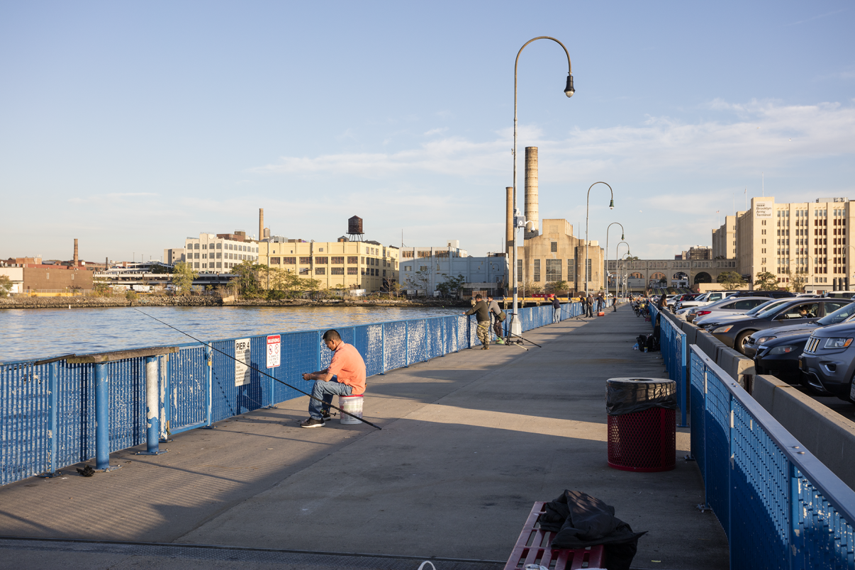 A person sits on a stool fishing on a blue metal pier under a clear sky. Nearby, people walk along the pier lined with vintage-style street lamps. Industrial buildings and parked cars can be seen in the background.