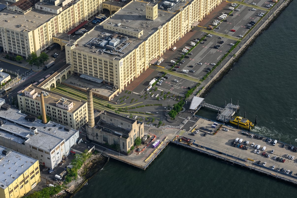 Aerial view of a coastal industrial complex with large warehouse buildings, parking lots, and a dock. A waterfront area with a docked yellow vessel on the right and surrounding streets and green spaces is visible.