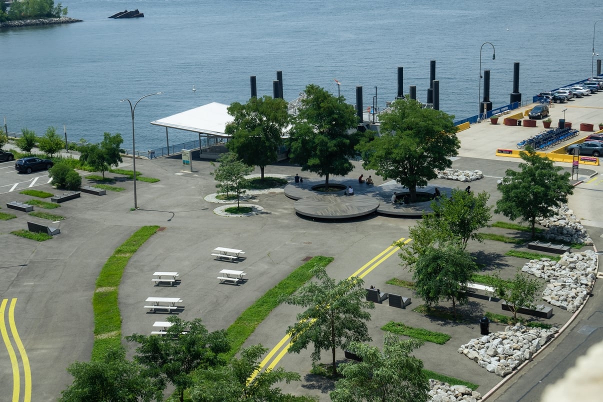 Aerial view of an empty waterfront park with curved pathways, picnic tables, rock formations, and trees. A circular seating area is in the center, while a dock extends into the water. Cars are parked nearby under a clear sky.