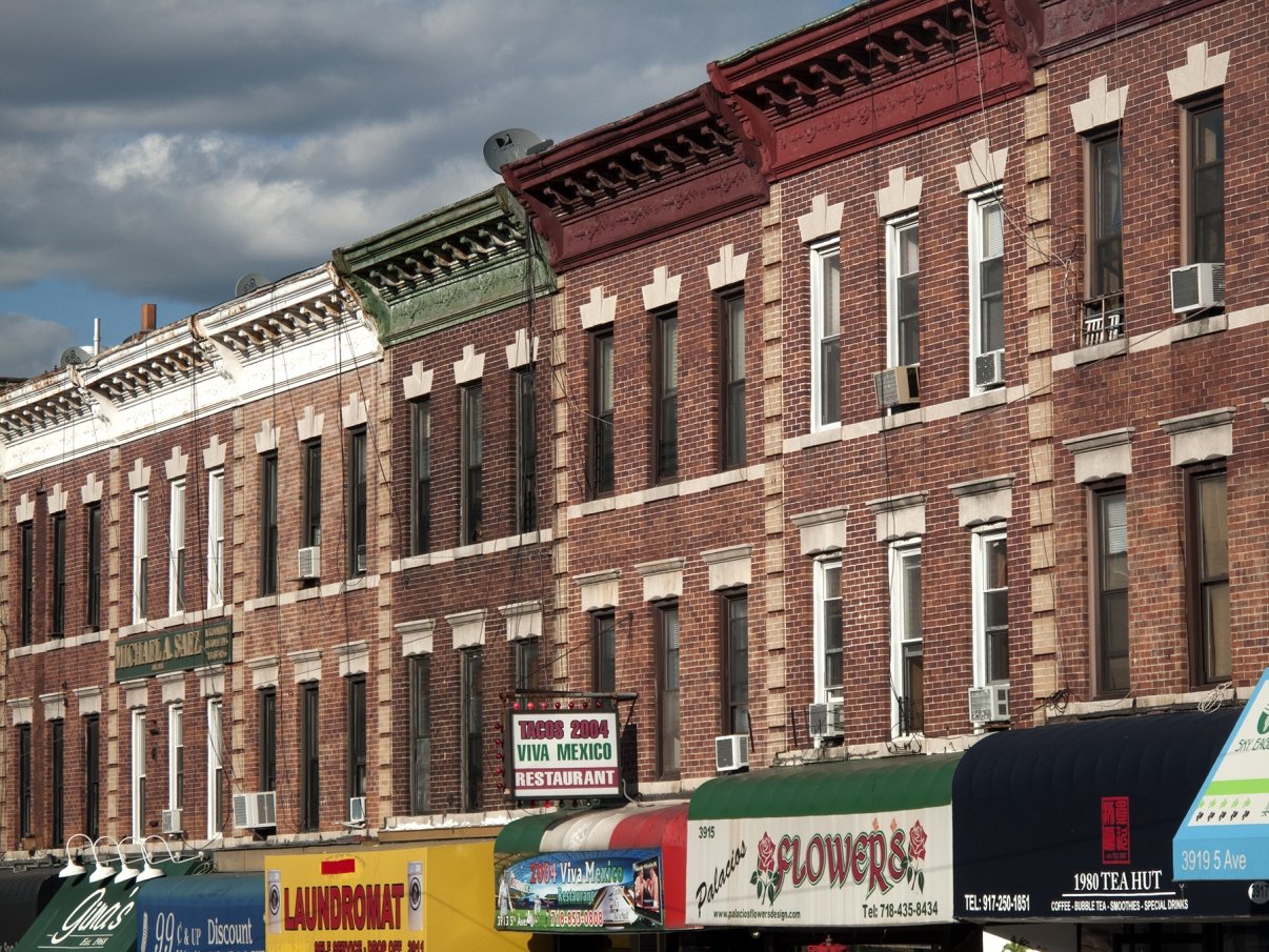 A row of colorful brick buildings with various storefronts below, including a flower shop and a restaurant named Viva Mexico. The sky is partly cloudy, creating a shadowed effect on the buildings.