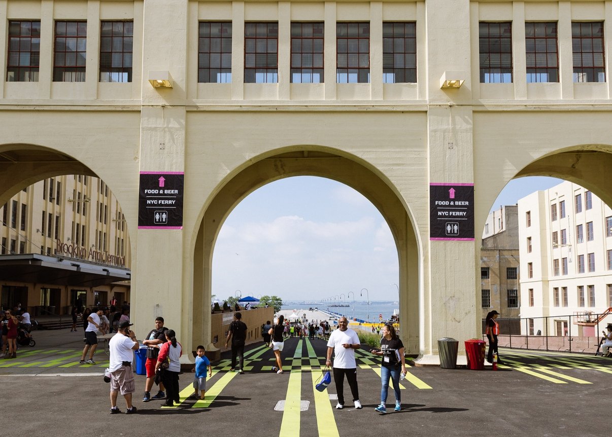 People walk under large arches at Brooklyn Army Terminal. The building has a pale exterior with windows above. Signs direct to food, beer, and the NYC ferry, and the water is visible in the background. The ground is marked with yellow lines.