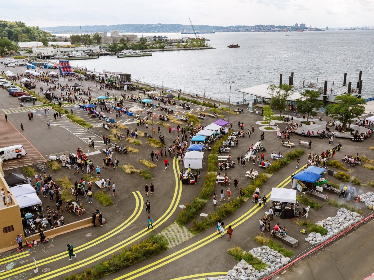 Aerial view of an outdoor food festival by the waterfront. Crowds of people gather around various food stalls and seating areas. The scene includes colorful tents and a view of the water with ships in the distance.