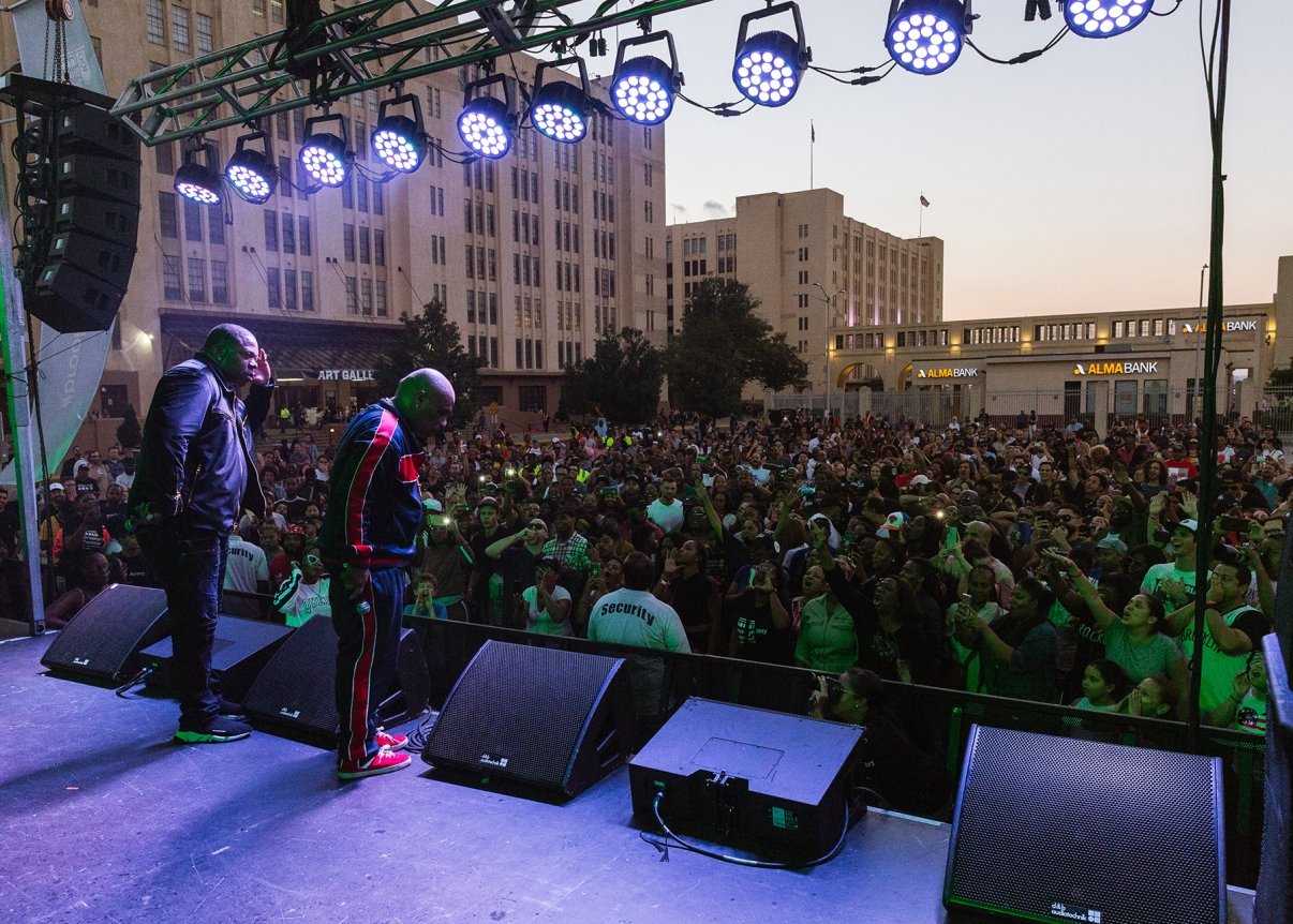 Two performers stand on an outdoor stage, one in a tracksuit. A large, enthusiastic crowd is gathered in front. Stage lights overhead illuminate the scene. A cityscape with tall buildings is visible in the background.