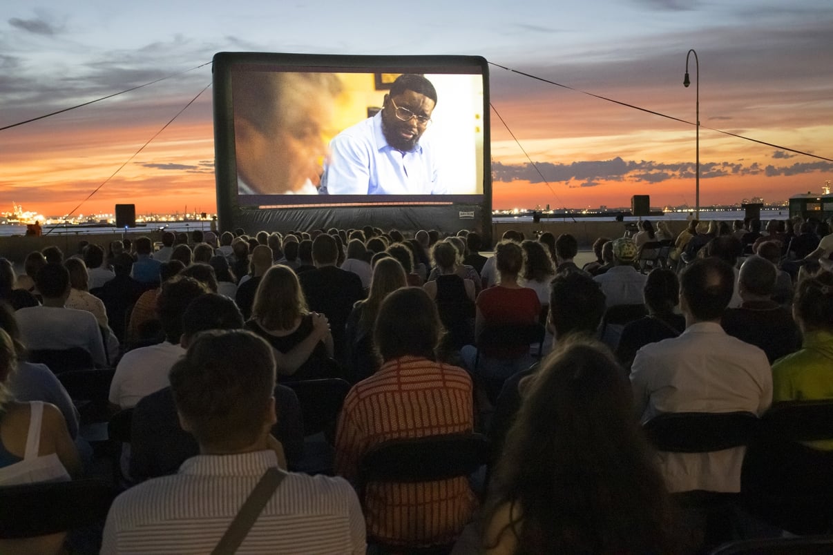 A large crowd watches an outdoor movie on a big screen at sunset. The sky is a vibrant mix of orange and blue hues, creating a picturesque backdrop. The audience sits in rows of chairs facing the screen.