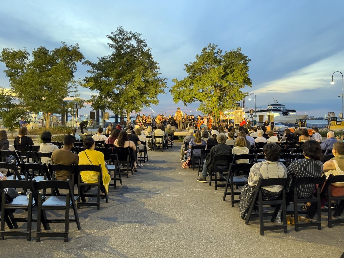 An outdoor orchestra performance in a park at sunset, with rows of seated audience members watching. Trees and a waterfront view in the background, along with a docked ferry. The sky is cloudy with a warm glow from the setting sun.