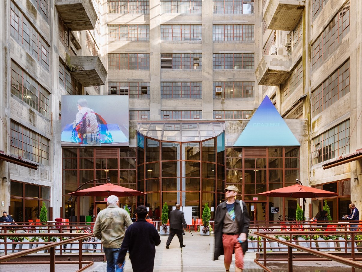 People walk through an industrial courtyard with tall concrete buildings. A glass entrance is flanked by colorful signage and red umbrellas cover outdoor seating. Large windows and art installations are visible on the building facades.