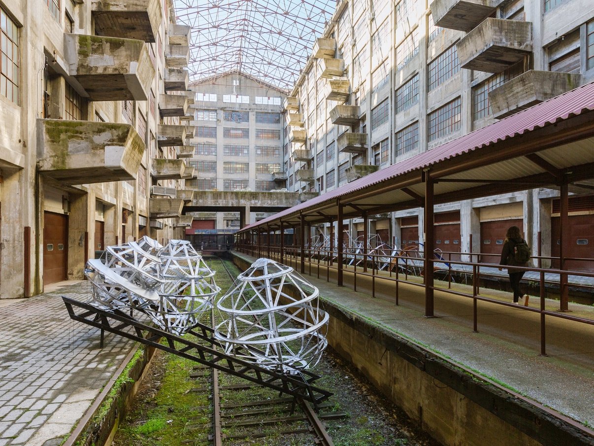 An abandoned industrial building features large geometric metal sculptures along old railway tracks. The structure has an open roof with glass panels, and a person is walking on a parallel pathway under a red canopy.