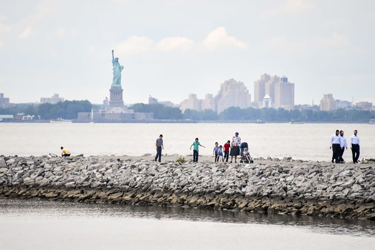 People walk along a rocky shoreline with the Statue of Liberty visible in the background across the water. A few individuals are clustered together, and the sky is cloudy. Nearby buildings can be seen on the horizon.