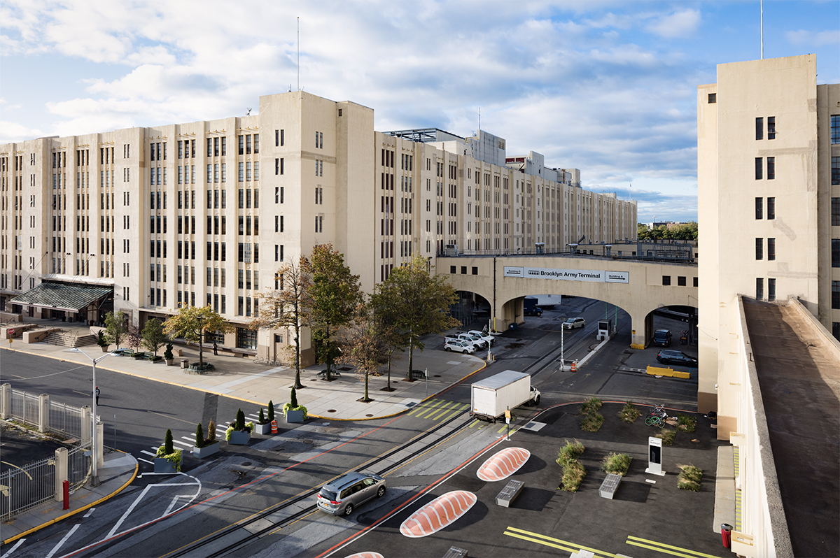 A large Art Deco-style building with multiple stories and tall, narrow windows. A roadway passes beneath an arched bridge that connects the building sections. Trees and parked cars are visible, and the sky is partly cloudy.